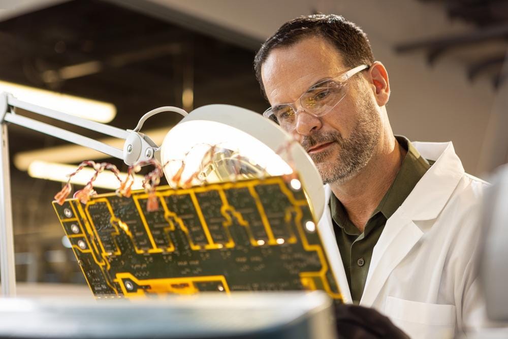 Un ingeniero con bata de laboratorio y gafas de seguridad inspecciona una placa de circuito bajo una lámpara de aumento.