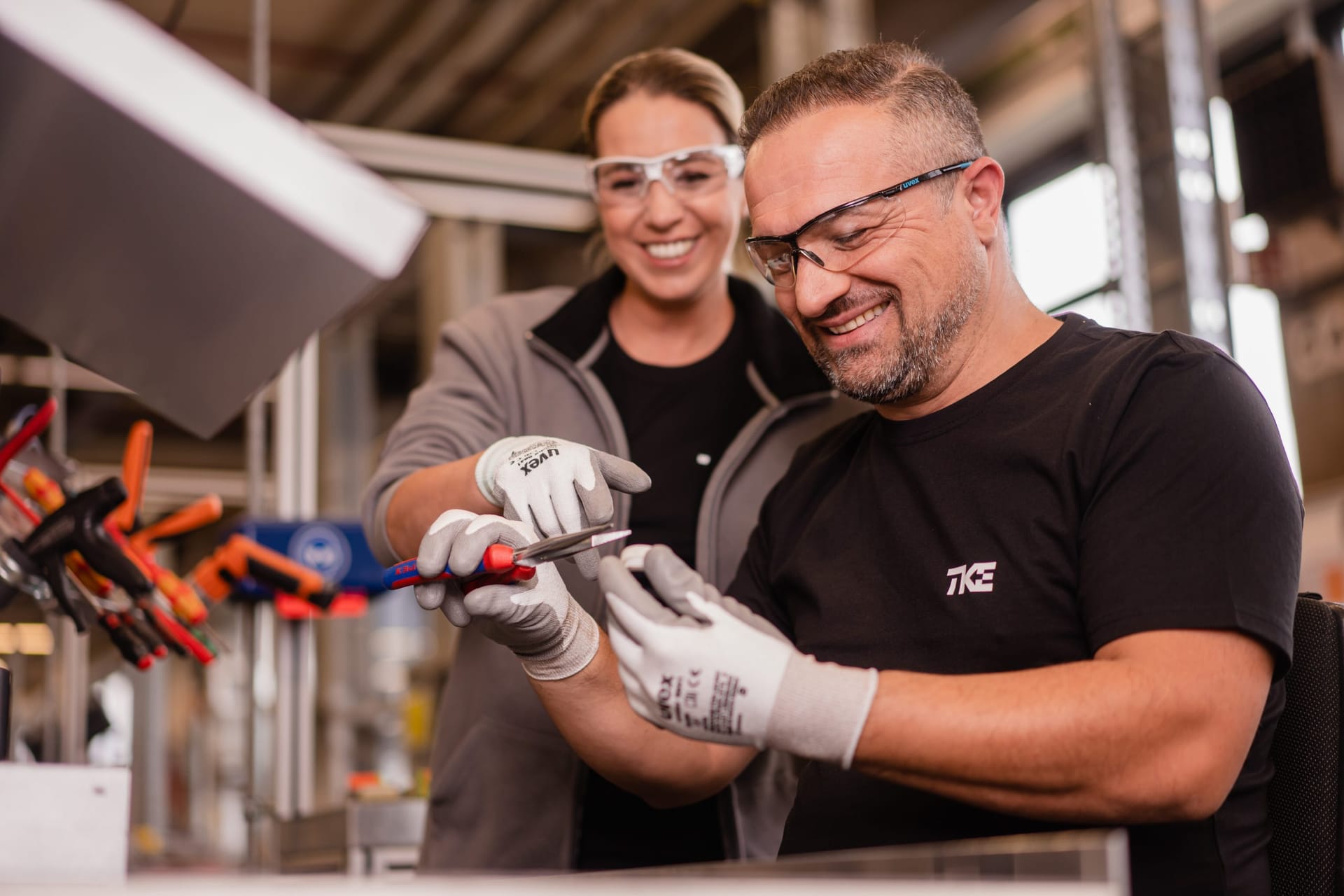 Dos compañeros sonrientes con gafas de seguridad y guantes de trabajo manipulan juntos una herramienta en un banco de trabajo.