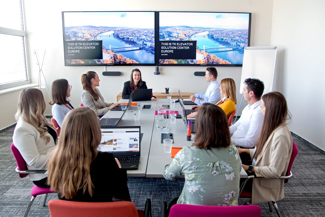 Un equipo se reúne en torno a una larga mesa de conferencias bajo dos pantallas con el título «This is TK Elevator Solution Center Europe».