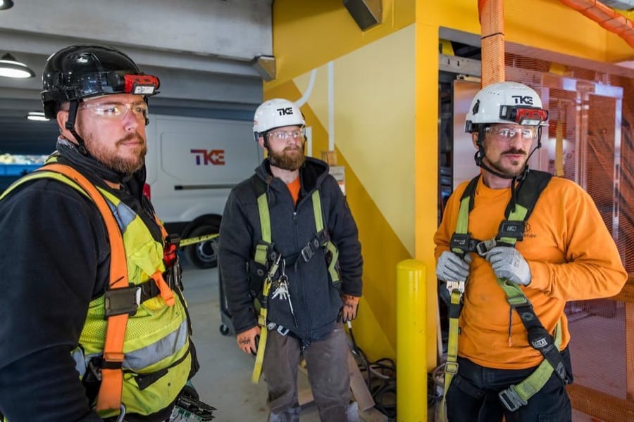 Tres técnicos de TKE con casco, gafas de seguridad y arnés en una obra.